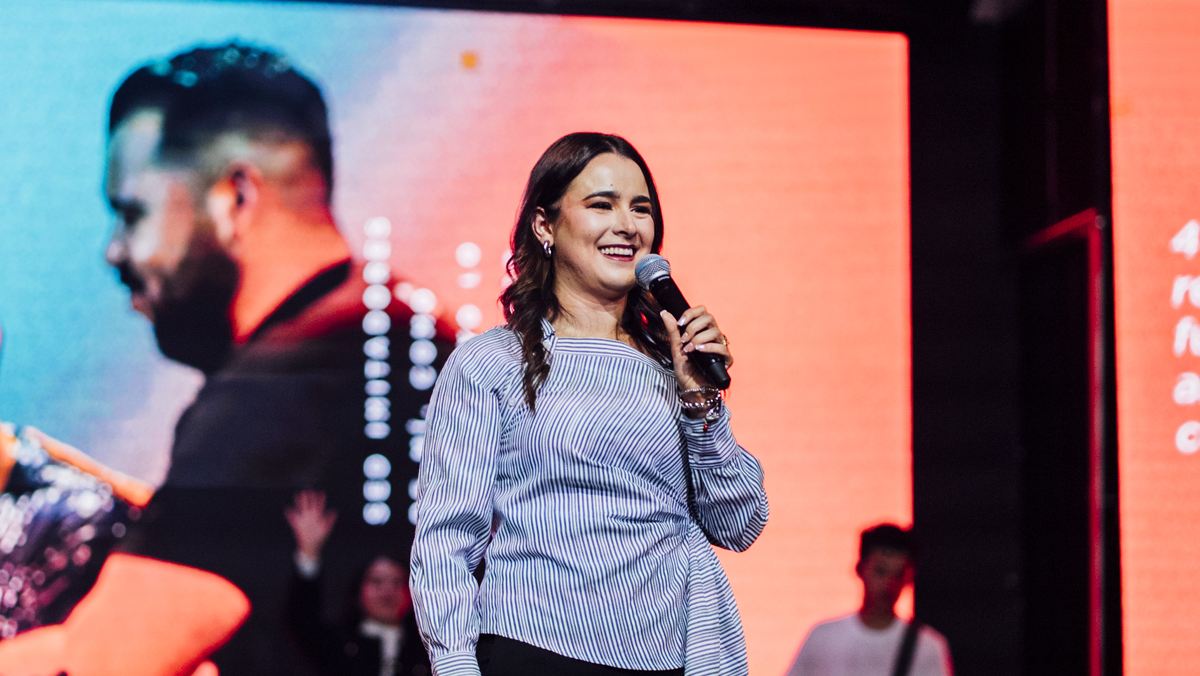 Michelle Ruiz con micrófono frente a una pantalla roja, sonriendo mientras habla.